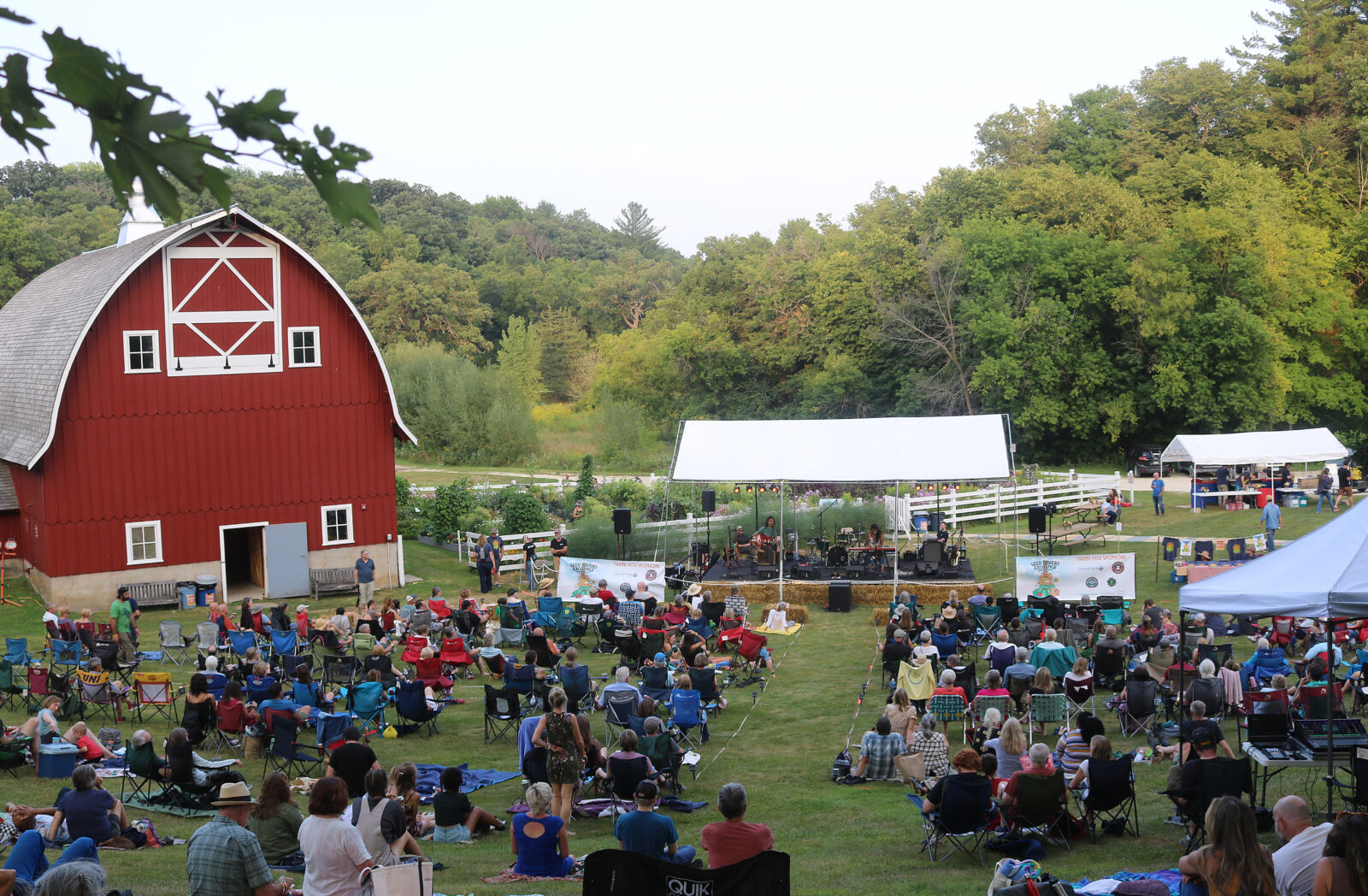 A crowd gathered on a lawn in front of a stage and red barn.