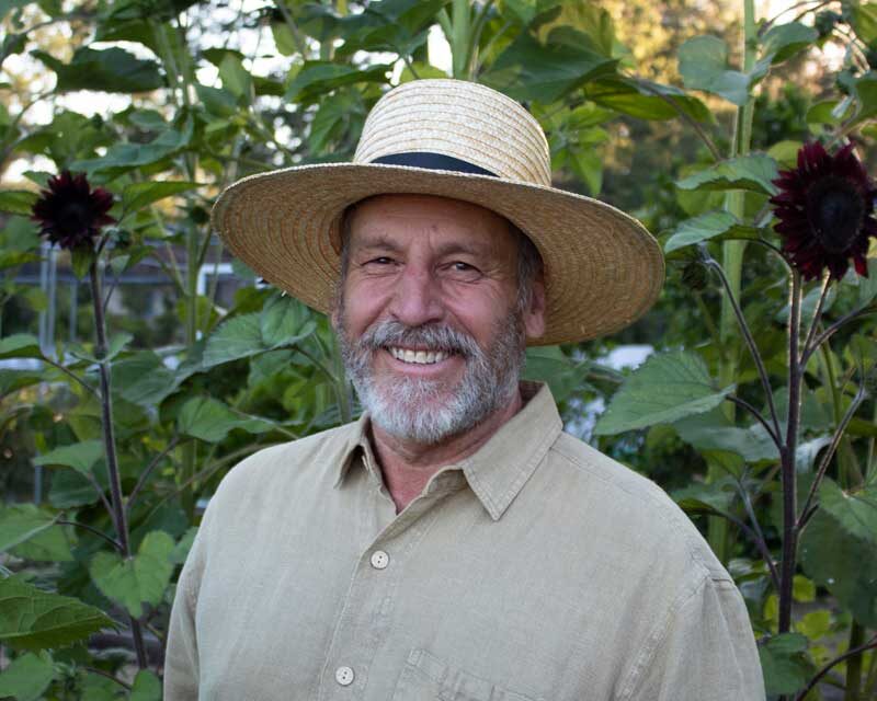 Man with a beard in a wide-brimmed hat and khaki button-down shirt smiling at the camera.