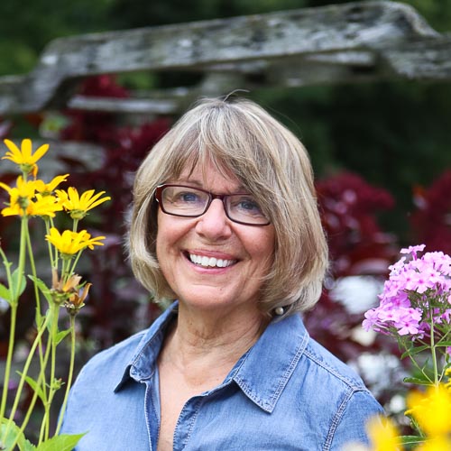 Woman smiling in glasses and a blue button-down shirt surrounded by flowers.