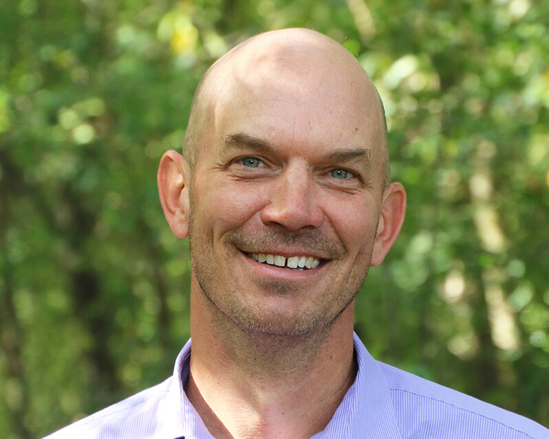 Man wearing a pale purple button-down shirt smiles into camera.