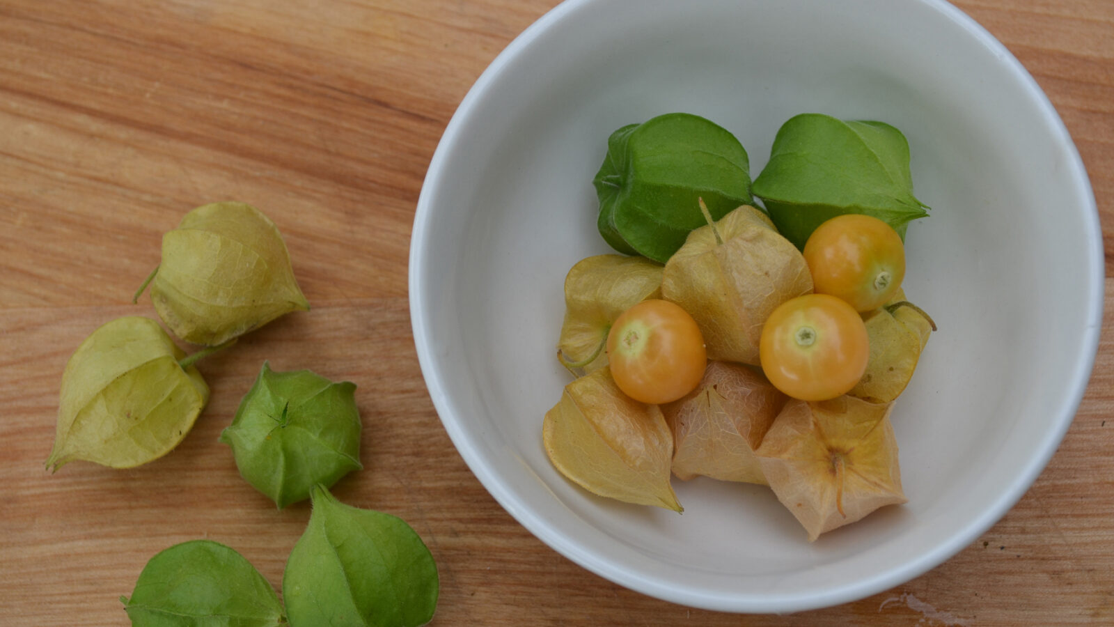 green and yellow ground cherries in a white bowl and on a wooden surface
