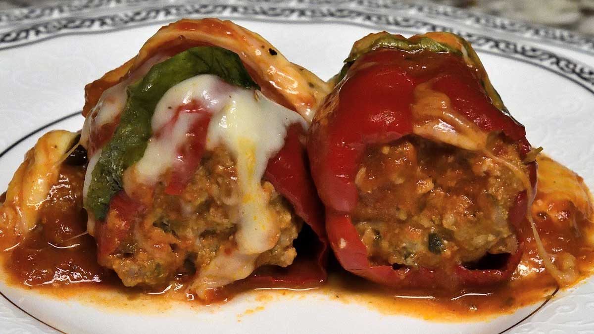 Two pieces of cooked ground meat stuffed inside of two red peppers, with cheese, small green leaves, and sauce on top