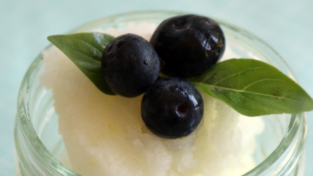 A glass jar with pale yellow sorbet topped with blueberries and small green leaves