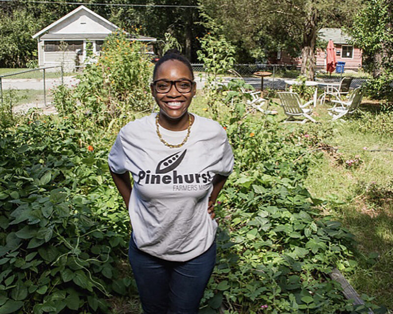 Black woman in a grey t-shirt stands proudly smiling to camera in front of a small garden.