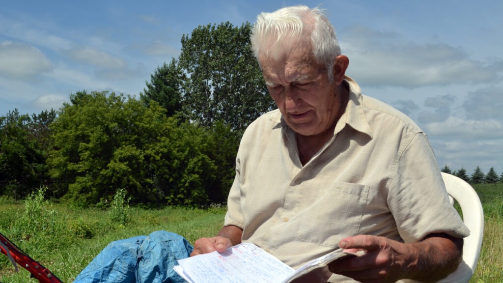 A man in a tan shirt sits and reads a notebook outside