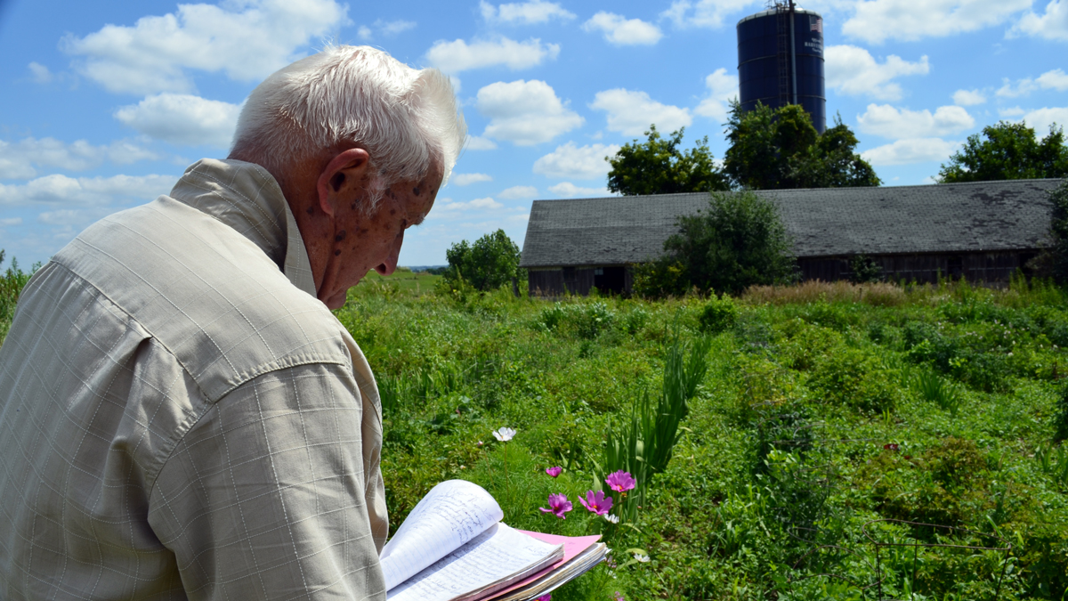 A man holds a notebook while looking over a garden, with a barn in the background