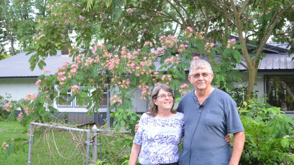 A man and woman stand in front of a tree with pink blossoms, a wire fence, and a house