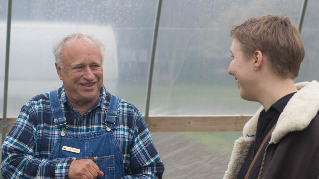 A man in overalls and a plaid shirt smiles at another person wearing a coat in a greenhouse