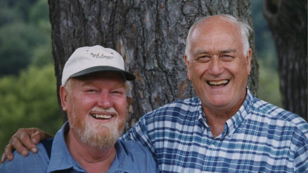 A man in a blue shirt and white baseball cap and a man in a blue plaid shirt smiling together in front of a tree