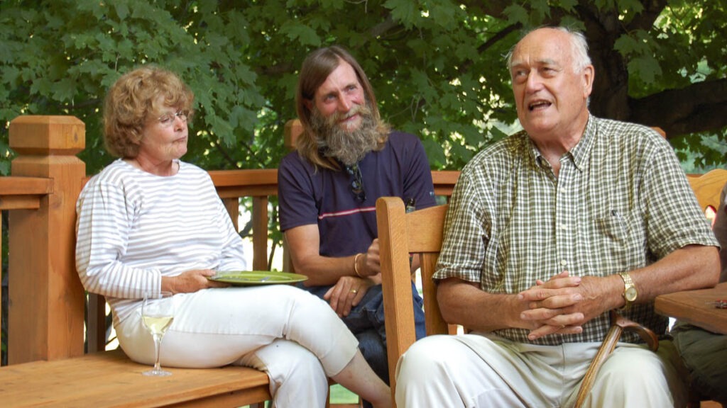 A man speaking sits in a chair while a woman and a man watch while sitting on a bench outdoors