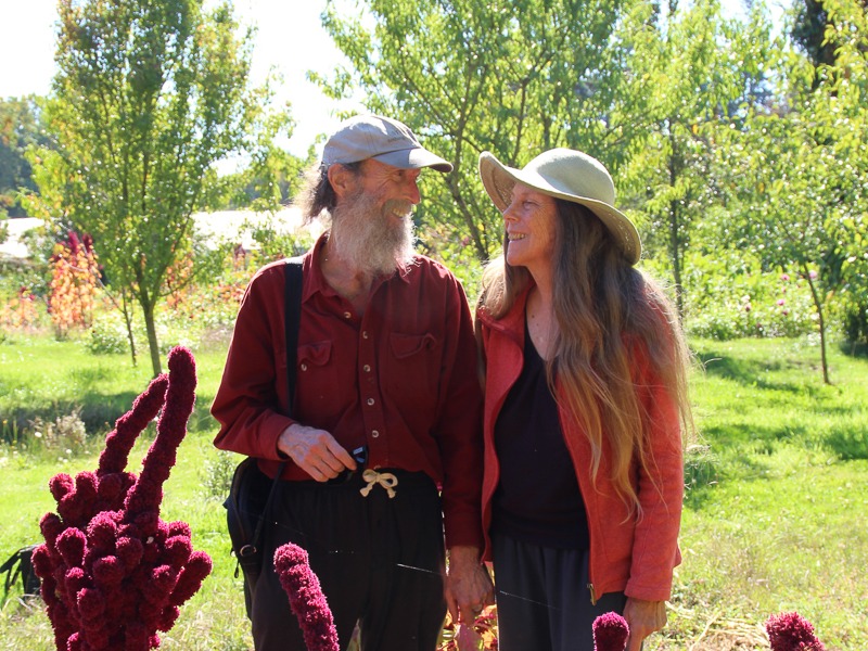 Man with a long white beard in a baseball cap and red button-down shirt smiling to the woman in a wide-brimmed hat looking back at him. Both are smiling.