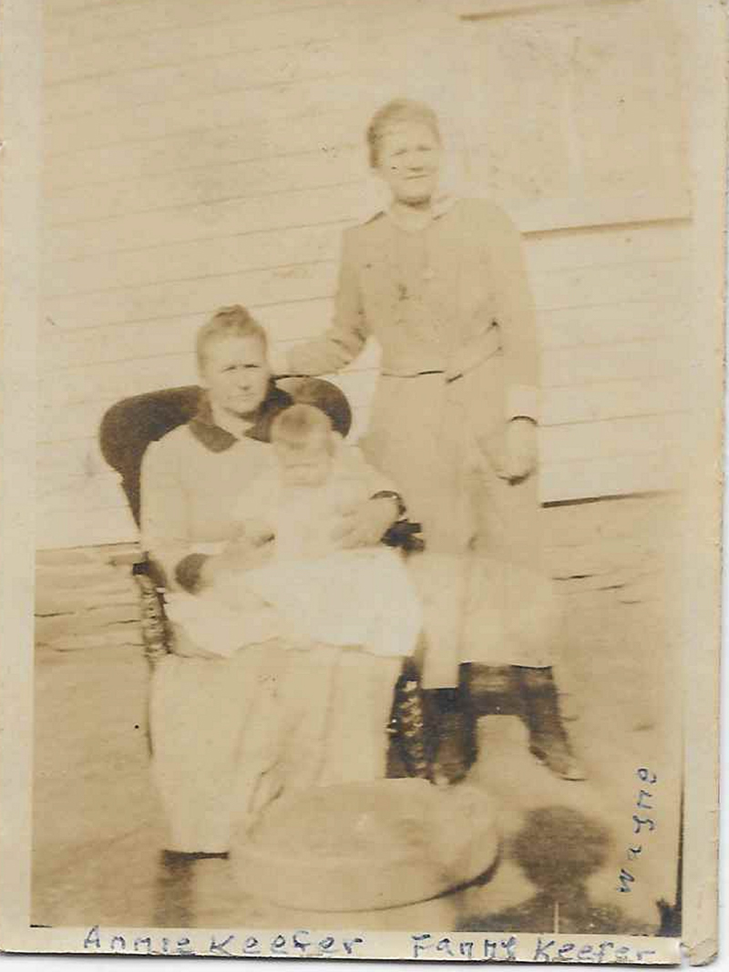 An old sepia photo of a woman holding a baby in a chair, while another woman stands behind her