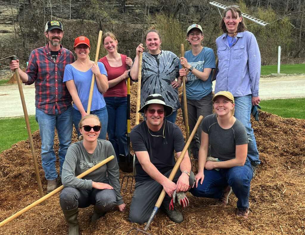 Group of people pose facing the camera on a pile of mulch holding garden equipment.