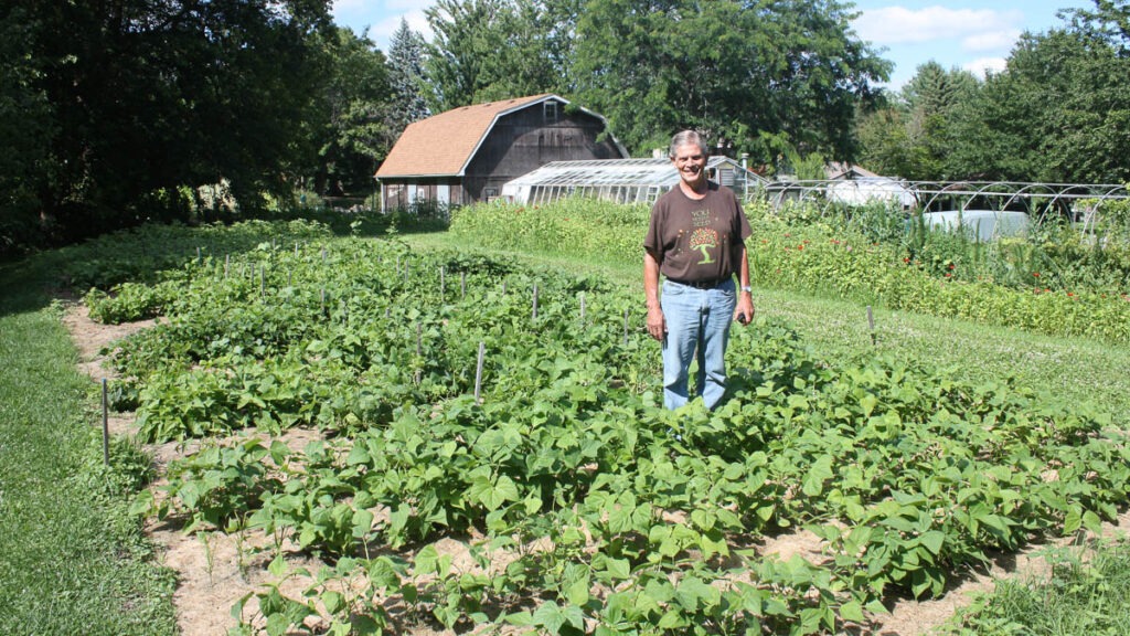 A man stands in a garden with rows of plants, with a barn and greenhouse in the background