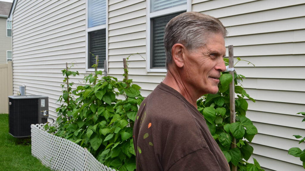 A man stands next to a garden bed with plants growing up tall wooden stakes, next to the side of a house