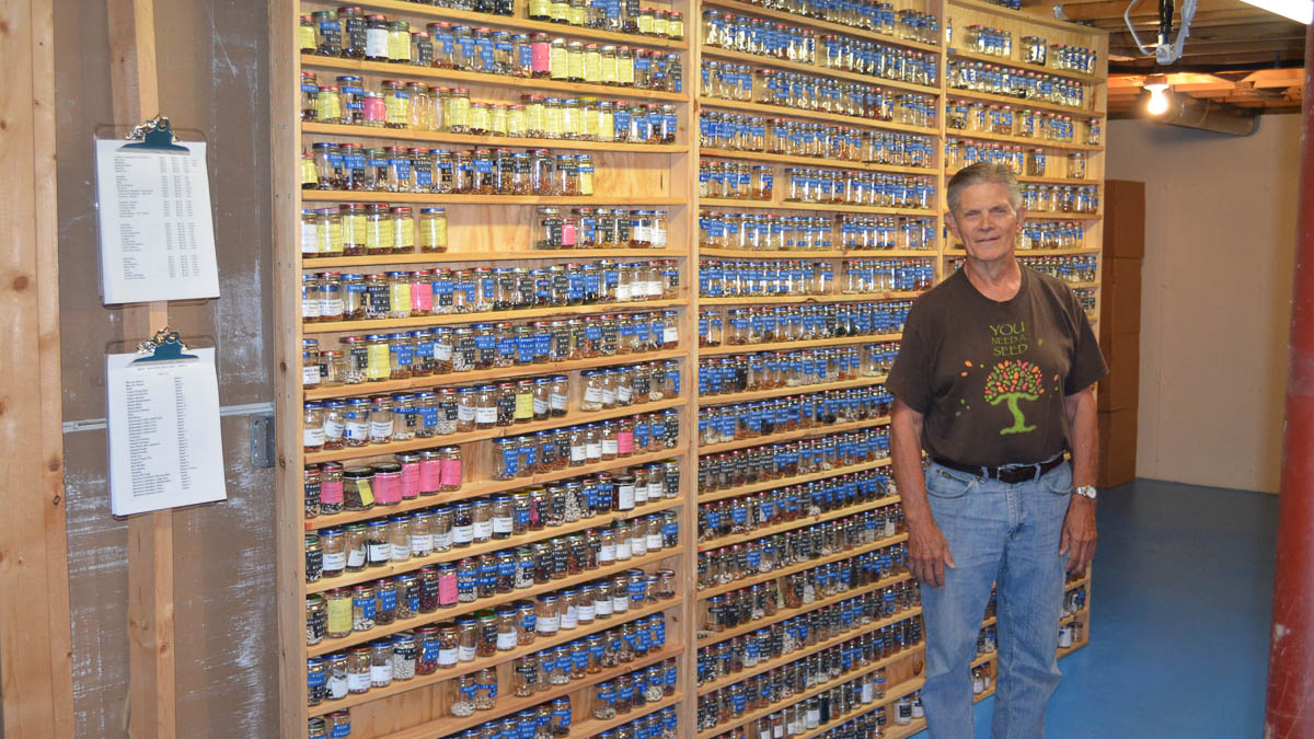 A man, Russ Crow, stands in front of tall shelves with many small jars of seeds