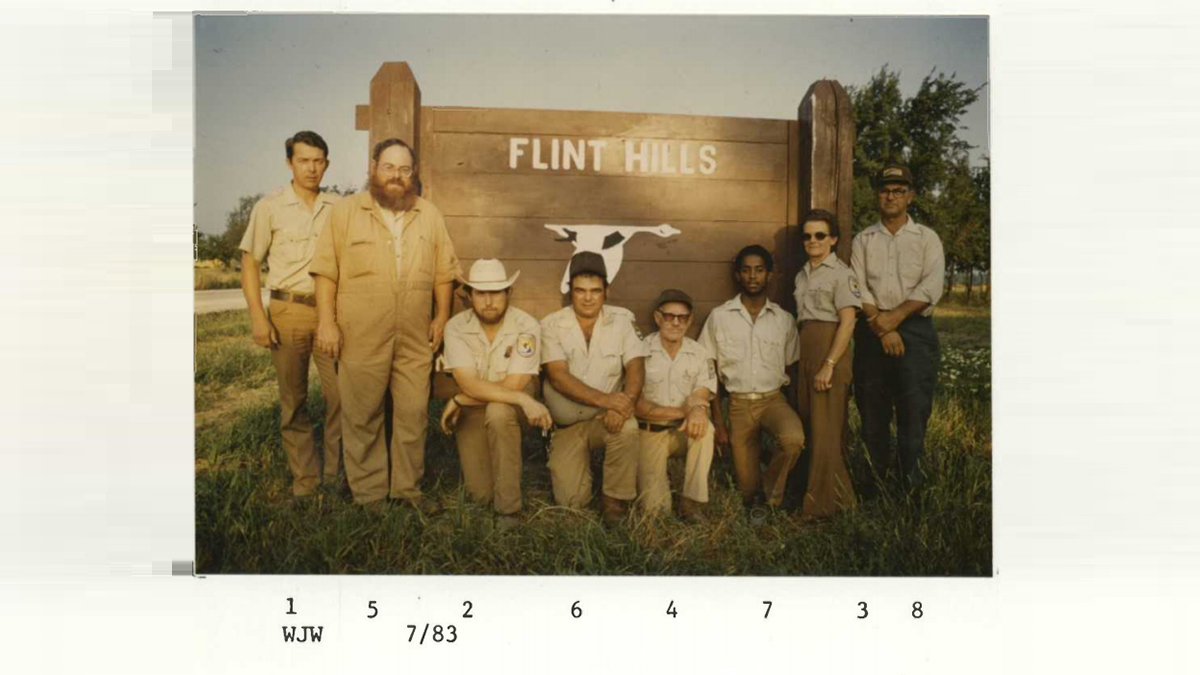 Eight people in tan uniforms pose in front of a wooden "Flint Hills" sign