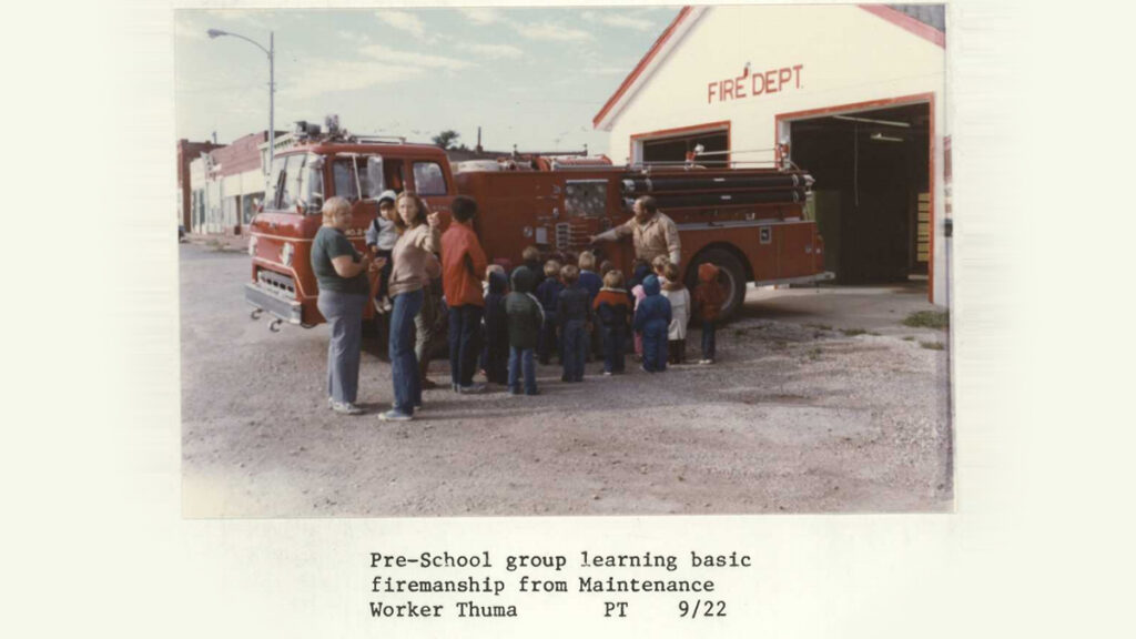 A group of adults and children gather next to a firetruck outside of a firehouse garage