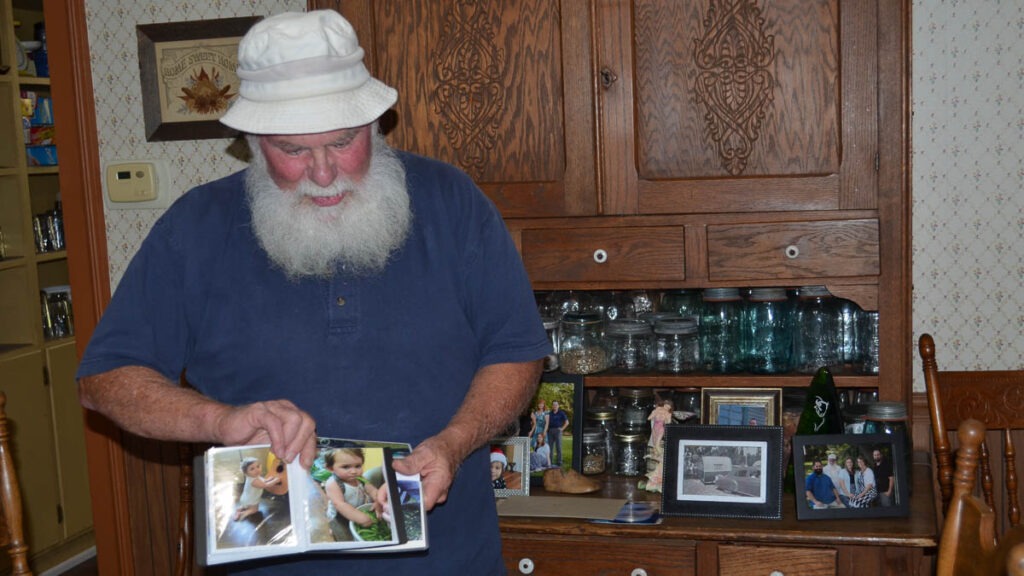 A man holds a photo album next to a cabinet with glass jars and picture frames on it