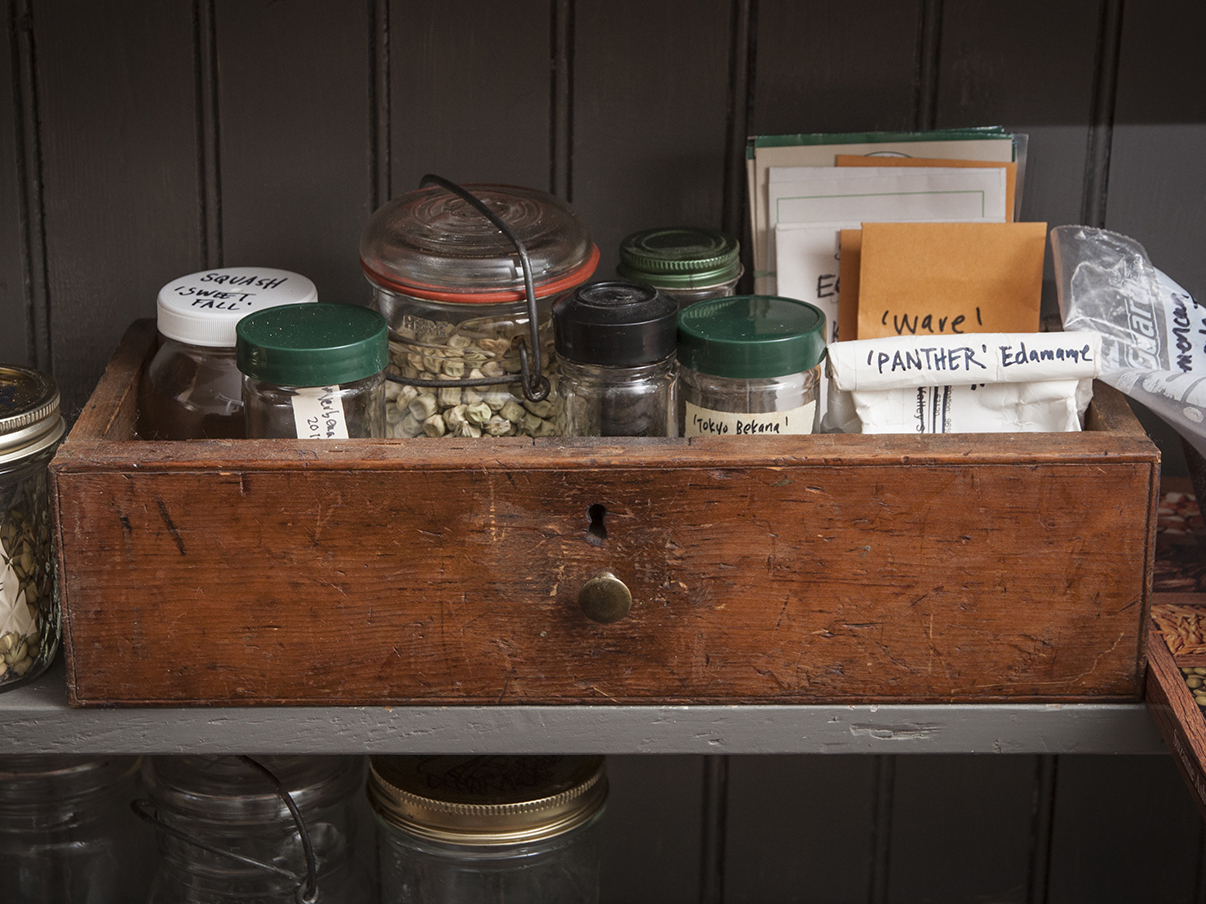 A wooden box with jars and packets of seeds inside sits on a shelf