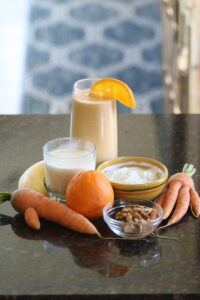 An orange smoothie on a table with carrots, an orange, a glass of milk, a banana, nuts, and a bowl of yogurt