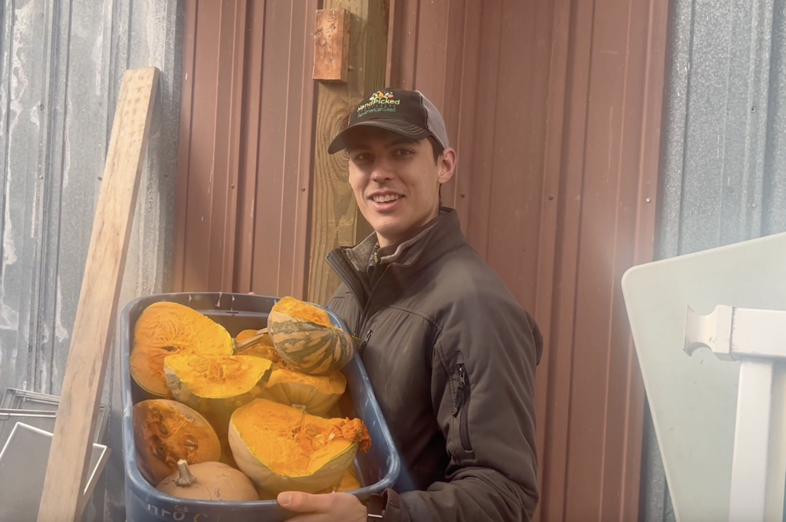 A man in a baseball cap smiles and holds up a large bucket with cut up squash.