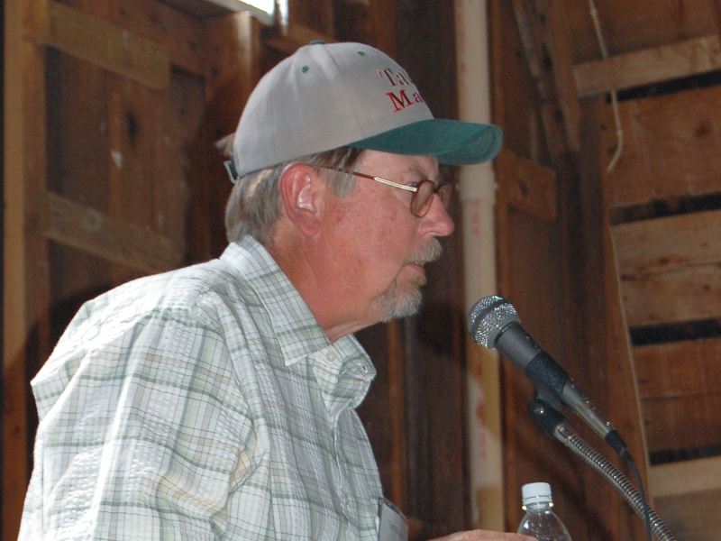 A man wearing a plaid shirt and a baseball cap speaks into a microphone in a barn