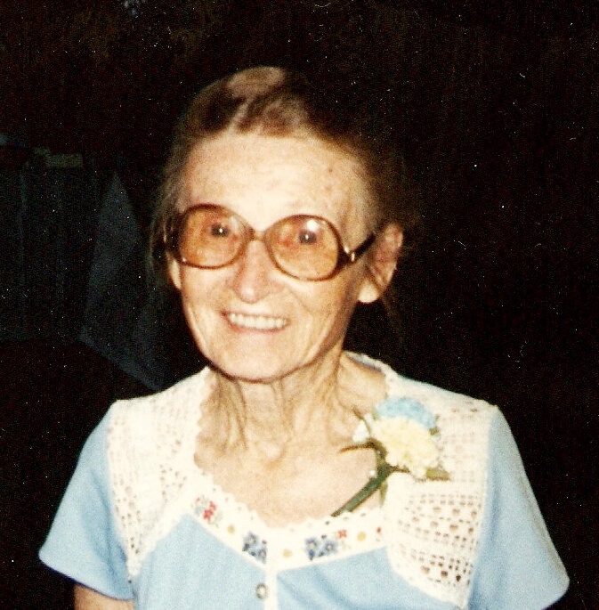 An older woman smiling, wearing glasses and a blue dress with flower pinned to it.