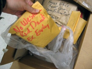 A hand pulls a yellow manila envelope from a box with other envelopes and a plastic bag.