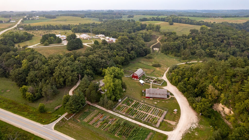 An aerial view of a farm with gardens, several buildings, a barn, trees, and fields