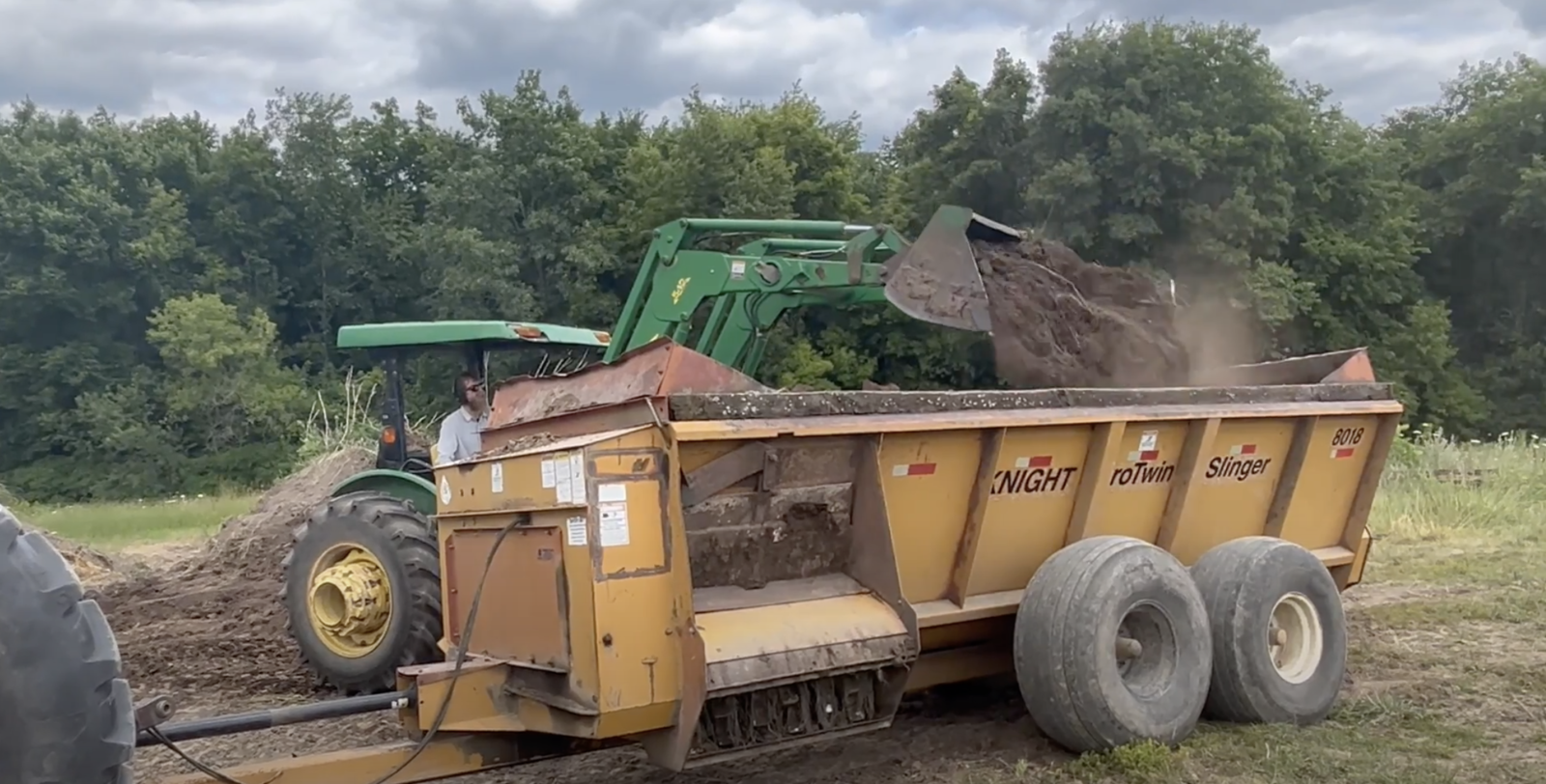 A green tractor dumps dirt-like compost into a large trailer