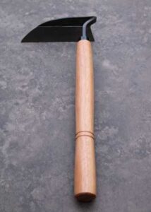A hand hoe garden tool with a dark gray head and wooden handle rests on a gray surface