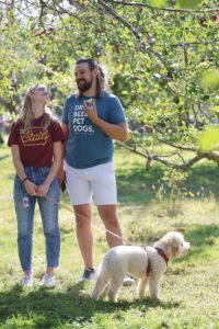 Two people stand in an apple orchard with a white dog.