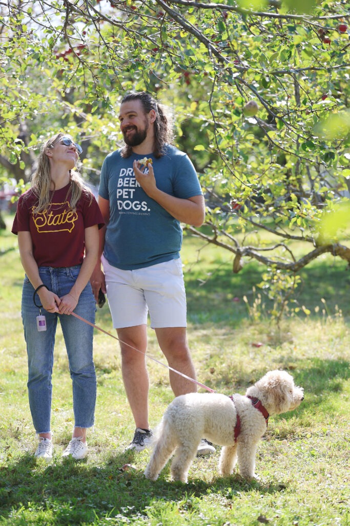 Two people stand in an apple orchard with a white dog.