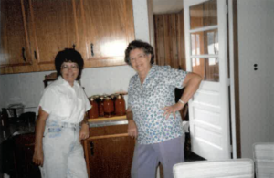 Two women pose standing in a kitchen in front of many cans of 'Oma's Orange' tomato cocktails