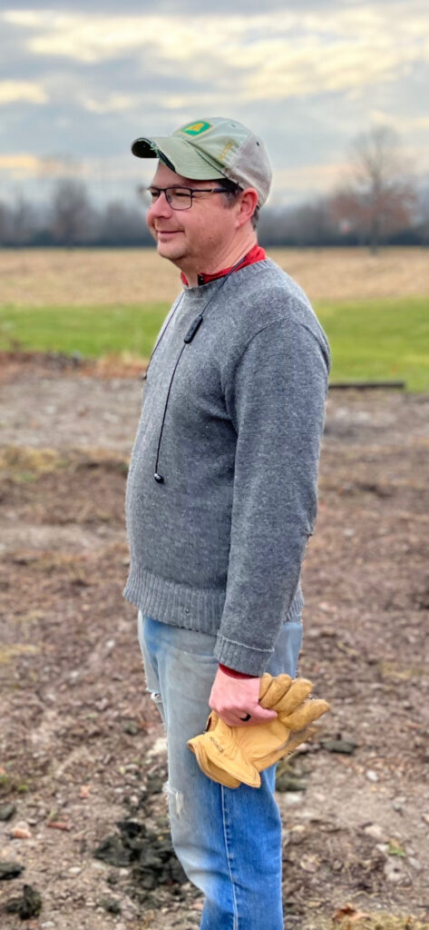 Man standing, holding gloves and wearing a baseball cap in a field.