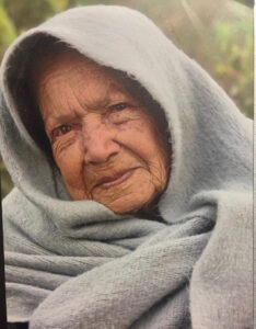 An older woman wearing a scarf wrapped around her head and shoulders looks into the camera