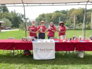 Four women holding jars of seeds stand behind a table outdoors