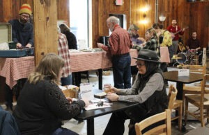 Two women sit at a table, with other people standing at long tables in the background