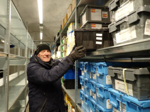 older man in black cap and black jacket placing black storage box on a shelf inside a storage facility 