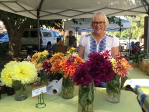 Woman in glasses standing under tent and behind a table with vases of dahlias in different colors. 