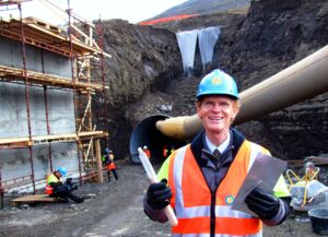 middle-aged man wearing light-blue hard had and orange safety vest stand in front of excavated building site