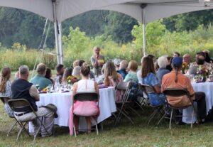 A woman addresses a crowd of people sitting at tables under a large tent