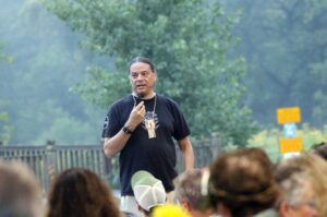 A man in a black t-shirt holds a microphone and addresses a crowd