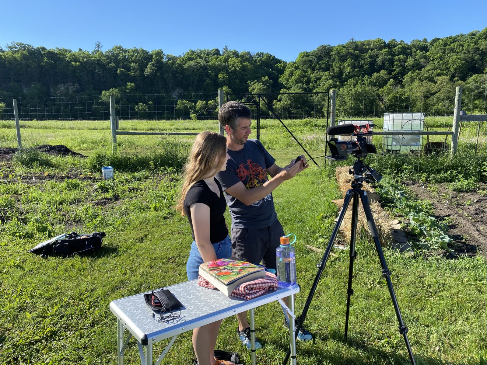 woman and man stand next to white table examining a camera on a tripod amid a garden