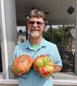 A man smiles in front of a window holding two very large tomatoes