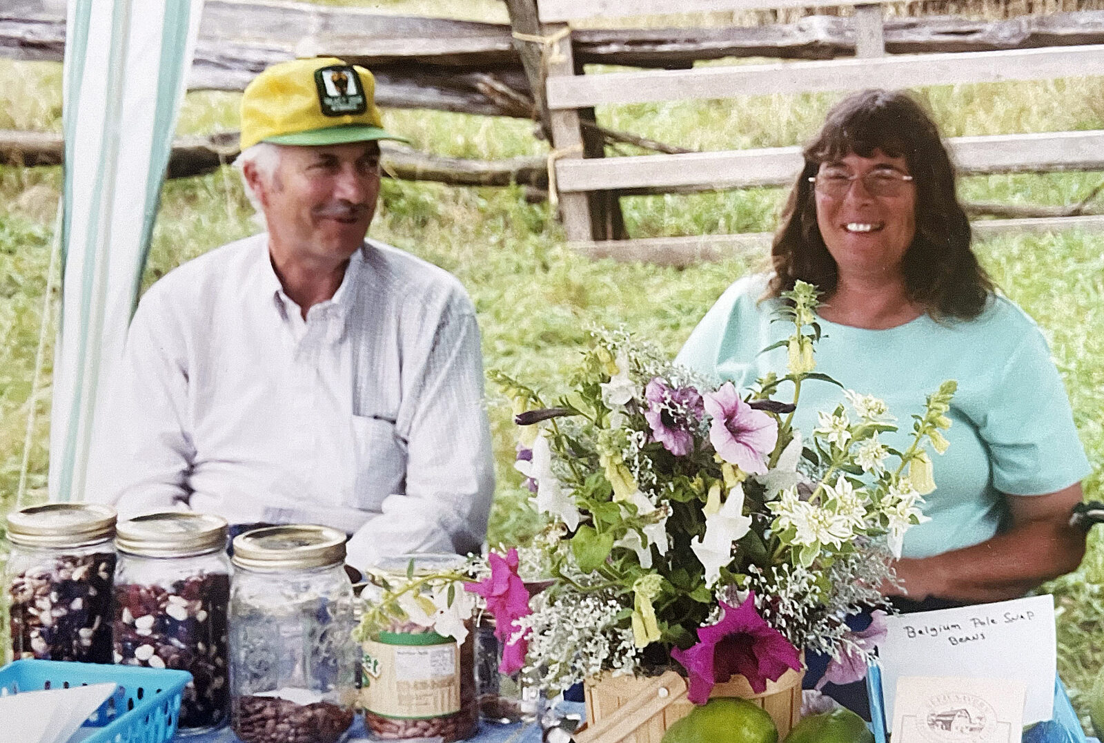 man and woman sitting outdoors behind a table displaying beans in jars and fresh flowers