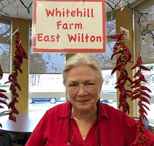 A woman in a red shirt smiles beneath a hanging sign that says "Whitehill Farm East Wilton" with strings of red peppers hanging on either side of her.