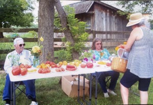 man and woman sit outdoors at long white table holding fresh produce while another woman stands at front, right 