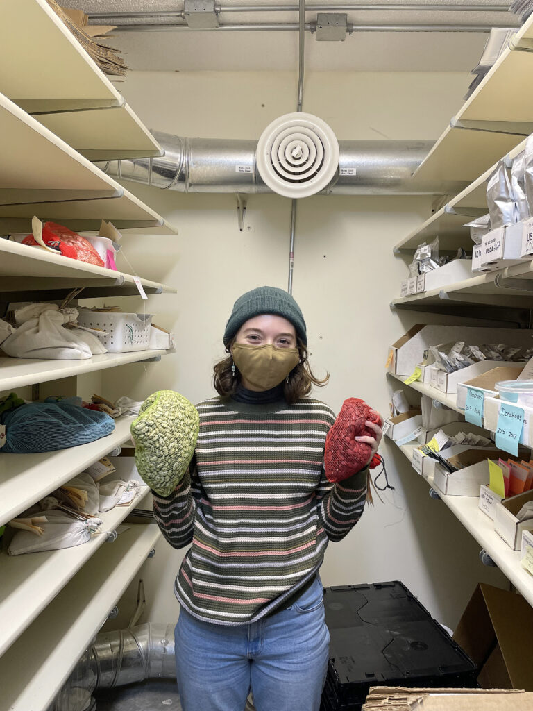 young woman wearing beanie and face mask, holding bags with seeds, flanked by shelves with seeds